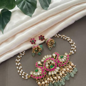 May include: A gold necklace with a pink and green floral design, featuring a pearl chain and dangling green stones. The necklace comes with matching earrings.