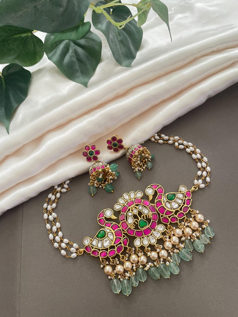 May include: A gold necklace with a pink and green floral design, featuring a pearl chain and dangling green stones. The necklace comes with matching earrings.
