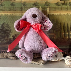 Puede incluir: Un juguete de peluche con forma de perro lavanda, con nariz marrón y orejas caídas. Tiene un lazo de satén rojo brillante y tela estampada floral en el interior de las orejas y las patas. El juguete está sobre madera flotante.