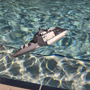 May include: A gray model warship featuring a sleek, angular hull design with a pointed bow, a central superstructure with radar masts, and a flat aft deck with a circular landing pad, floating on the surface of clear, rippling blue water.