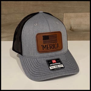 Può includere: Cappellino da camionista grigio con retro in rete nera e toppa in pelle marrone con bandiera americana e scritta 'Merica. Il cappellino ha una visiera curva e un cinturino regolabile.