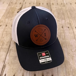 May include: Navy blue and white trucker cap with a brown leather patch. The patch features the letters "W" and "I" with crossed arrows and the text "Est. 1848". The cap has a curved peak and a Richardson 115 Low-Pro Trucker tag.