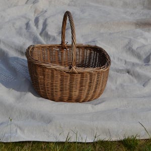 May include: A woven wicker basket with a handle. The basket is oval-shaped and made of light brown wicker. The handle is a twisted rope design. The basket is sitting on a white sheet.