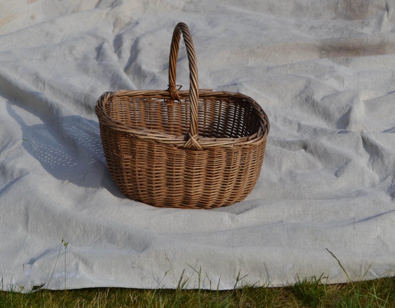 May include: A woven wicker basket with a handle. The basket is oval-shaped and made of light brown wicker. The handle is a twisted rope design. The basket is sitting on a white sheet.