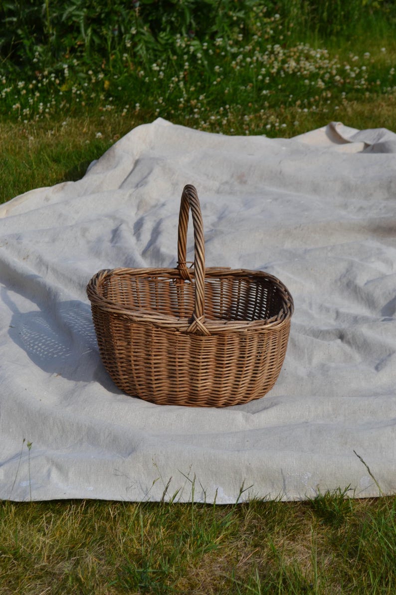 May include: A woven wicker basket with a handle. The basket is oval-shaped and made of light brown wicker. The handle is arched and made of twisted wicker. The basket is sitting on a white sheet on the grass.