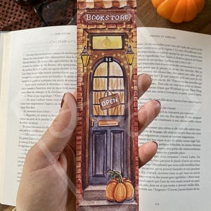 Peut inclure: Marque-page aquarelle représentant une façade de librairie avec une porte, une citrouille et le mot "OPEN". Le marque-page a un extérieur en briques, un auvent rayé et de petites lumières. Le design est posé sur un livre ouvert.