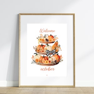 May include: A framed print featuring a watercolor illustration of a tiered tray filled with pumpkins, gourds, and fall foliage. The words "Welcome October" are written in a decorative font. The artwork is set against a white background.