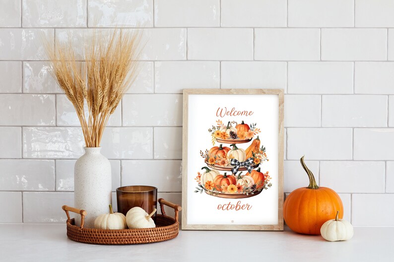 May include: A framed watercolor print with a tiered tray of pumpkins and the text "Welcome october." A vase of wheat, a decorative tray with pumpkins, and a candle are also present. An orange pumpkin and a white pumpkin are on the counter.
