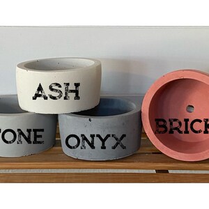 May include: Four small concrete planters in different colors. The planters are labeled "ASH", "STONE", "ONYX", and "BRICK".