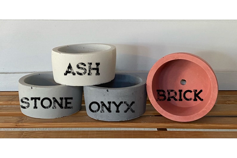 May include: Four small concrete planters in different colors. The planters are labeled "ASH", "STONE", "ONYX", and "BRICK".