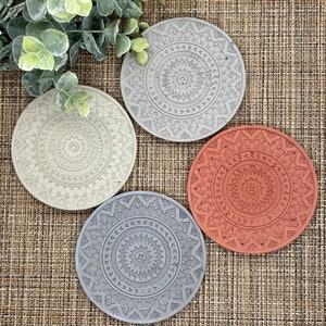 May include: Four round coasters with intricate mandala designs. The coasters are made of a light gray, dark gray, and terracotta material.