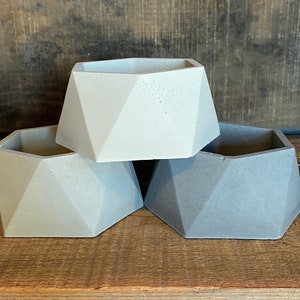 May include: Three geometric concrete planters in shades of gray. The planters are hexagonal in shape and have a rough, textured surface.