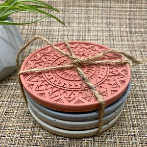 May include: Set of four round coasters with a geometric pattern. The top coaster is a terracotta color with a gray and white pattern. The coasters are tied together with twine.