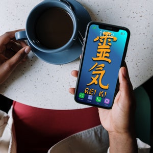 Può includere: Una persona tiene in mano uno smartphone con uno sfondo blu e la parola "REIKI" scritta in arancione. Lo schermo del telefono mostra anche i caratteri giapponesi per "Reiki" in giallo. La persona è seduta a un tavolo con una tazza di caffè.