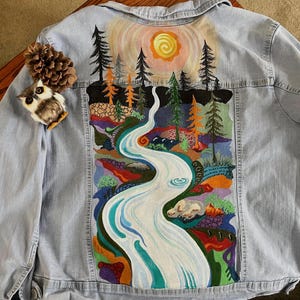 May include: Light blue denim jacket with a hand-painted landscape scene on the back. The artwork depicts a river flowing through a colorful forest under a bright sun. A small owl pin and pinecone are attached to the jacket.