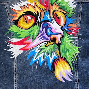 May include: A denim jacket with a colorful, abstract painting of a cat's face on the back. The cat's eyes are large and bright, and the fur is painted in a variety of colors, including green, blue, yellow, and pink.