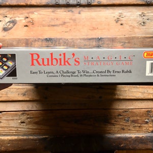 Pode incluir: Uma caixa cinzenta com uma imagem de um tabuleiro de jogo na frente. A caixa &eacute; etiquetada como "Rubik's MAGIC&trade; Strategy Game" e "Easy To Learn, A Challenge To Win...Created By Erno Rubik". A caixa tamb&eacute;m inclui o logotipo da Matchbox e um c&oacute;digo de barras.