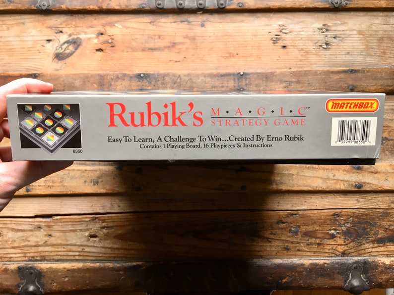 Pode incluir: Uma caixa cinzenta com uma imagem de um tabuleiro de jogo na frente. A caixa &eacute; etiquetada como "Rubik's MAGIC&trade; Strategy Game" e "Easy To Learn, A Challenge To Win...Created By Erno Rubik". A caixa tamb&eacute;m inclui o logotipo da Matchbox e um c&oacute;digo de barras.
