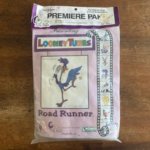 May include: A purple and white packaging for a "Premiere Pak" punch needle embroidery kit featuring Road Runner from Looney Tunes. The kit includes a pre-printed fabric, color coded yarn, and instructions. The packaging features a film strip design with images of various Looney Tunes characters.