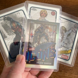 May include: Three Marvel trading cards in protective sleeves. The cards feature Iron Man, Silver Surfer, and Dagger. The Silver Surfer card is labeled "Marvel Icons". The cards have a metallic sheen and are set against a light brown background.