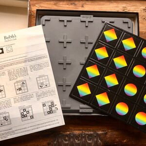 Pode incluir: Um jogo de estrat&eacute;gia da marca Rubik's com um tabuleiro de jogo preto e pe&ccedil;as de cores do arco-&iacute;ris. O tabuleiro de jogo tem um padr&atilde;o de grade e as pe&ccedil;as t&ecirc;m a forma de um c&iacute;rculo ou de um diamante.