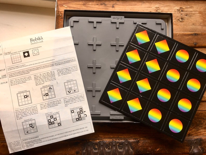 Pode incluir: Um jogo de estrat&eacute;gia da marca Rubik's com um tabuleiro de jogo preto e pe&ccedil;as de cores do arco-&iacute;ris. O tabuleiro de jogo tem um padr&atilde;o de grade e as pe&ccedil;as t&ecirc;m a forma de um c&iacute;rculo ou de um diamante.