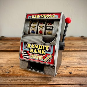 May include: A silver-colored metal slot machine coin bank labeled LAS VEGAS and BANDIT BANK JACKPOT. It features three spinning reels with symbols including stars, cherries, bars, and bells, and a black lever with a red ball handle on the side.