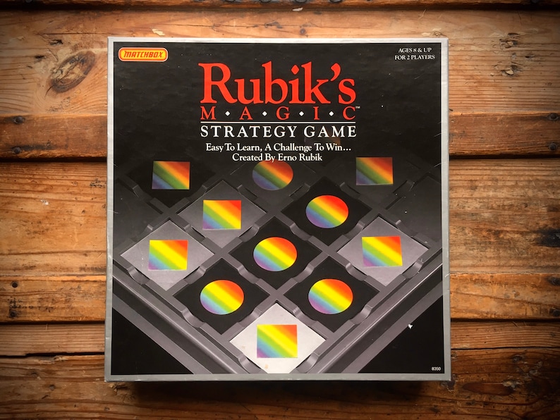 Pode incluir: Um jogo de tabuleiro em uma caixa preta com um design de arco-&iacute;ris. A caixa apresenta o texto "Rubik's MAGIC STRATEGY GAME" e "Easy To Learn, A Challenge To Win... Created By Erno Rubik".