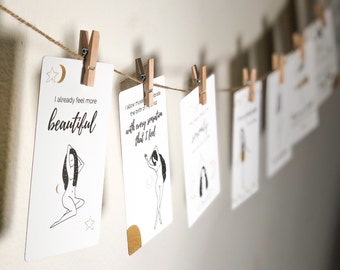 Puede incluir: Una cuerda de tarjetas con afirmaciones e ilustraciones de mujeres colgadas en una pared con pinzas para la ropa. Las tarjetas incluyen el texto "Ya me siento más hermosa" y "Me permito celebrar el nacimiento de mi lado salvaje con cada sensación que siento."