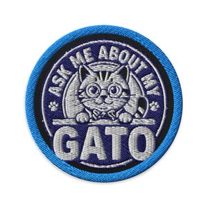 May include: A circular embroidered patch with a bright blue border. The center features a dark blue background with a white cat wearing glasses and a bow tie. The text reads ASK ME ABOUT MY GATO in white capital letters with two paw prints.