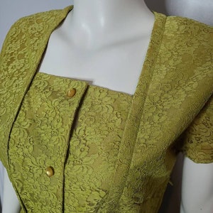 May include: A vintage-style, olive green dress with a square neckline and short sleeves. The dress features a delicate lace overlay and is adorned with gold-colored buttons down the front. The dress is displayed on a mannequin.