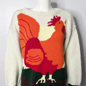 Peut inclure: Un pull en tricot à manches longues de couleur crème arborant un grand motif intarsia coloré représentant un coq. Le coq est illustré dans des tons orange vif, rouge et jaune, posé sur une base vert foncé. Le pull possède un col rond côtelé.