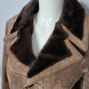 Puede incluir: Chaqueta de ante marrón claro con cuello y ribete de piel sintética marrón oscuro. La chaqueta tiene un diseño cruzado y detalles de costura visibles. El cuello es ancho y la chaqueta parece vintage.