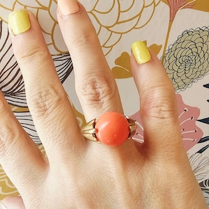 Peut inclure: Une bague dorée ornée d'une grosse pierre ronde de couleur corail. La bague a un anneau détaillé et texturé avec des accents en forme de feuilles de chaque côté de la pierre. La bague est portée sur une main avec des ongles peints.