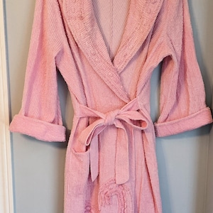Peut inclure: Un peignoir en tissu éponge rose clair doté d'un col châle, de manches longues à revers et d'une ceinture à nouer assortie. Le vêtement présente une surface en tissu côtelé texturé et un grand motif cachemire en relief sur le bas du devant.