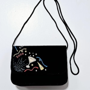 May include: Black rectangular shoulder bag with a long black cord strap. The bag features an embroidered design with the year "2000", a martini glass, confetti, and a party horn in gold, red, and blue.