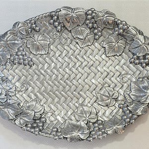 Puede incluir: Bandeja ovalada plateada con diseño de cesta tejida. La bandeja está decorada con hojas de uva y racimos de uvas en los bordes y tiene dos asas en forma de cuerda.