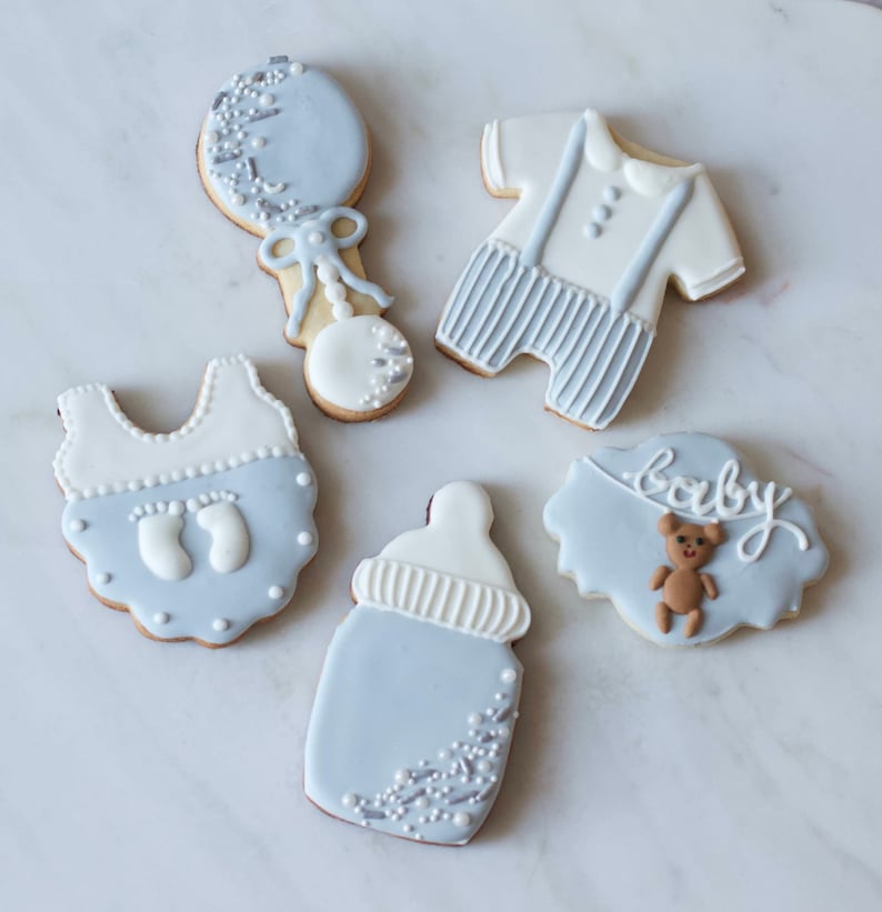 Puede incluir: Cinco galletas de az&uacute;car decoradas en azul y blanco. Las galletas tienen forma de biber&oacute;n, sonajero, body de beb&eacute;, atuendo de beb&eacute; con tirantes y una pancarta que dice "beb&eacute;" con un osito de peluche.