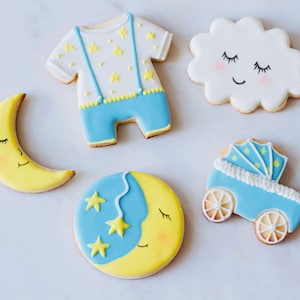 Puede incluir: Cinco galletas de az&uacute;car decoradas con un tema de beb&eacute;. Las galletas est&aacute;n decoradas con glaseado azul, amarillo y blanco. Una galleta tiene forma de media luna con una cara dormida, otra tiene forma de nube con una cara dormida, otra tiene forma de beb&eacute; en un mono con estrellas, otra tiene forma de cochecito con estrellas, y la &uacute;ltima es una galleta redonda con una media luna y estrellas.