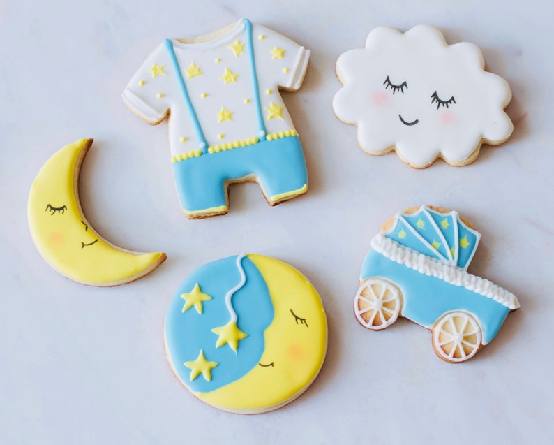 Puede incluir: Cinco galletas de az&uacute;car decoradas con un tema de beb&eacute;. Las galletas est&aacute;n decoradas con glaseado azul, amarillo y blanco. Una galleta tiene forma de media luna con una cara dormida, otra tiene forma de nube con una cara dormida, otra tiene forma de beb&eacute; en un mono con estrellas, otra tiene forma de cochecito con estrellas, y la &uacute;ltima es una galleta redonda con una media luna y estrellas.