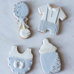 Puede incluir: Cuatro galletas de az&uacute;car decoradas con forma de biber&oacute;n, un body de beb&eacute;, un sonajero y un atuendo de beb&eacute; con tirantes. Las galletas est&aacute;n decoradas con glaseado blanco y azul claro y espolvoreadas con brillantina plateada.
