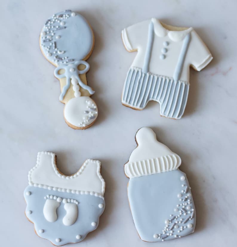 Puede incluir: Cuatro galletas de az&uacute;car decoradas con forma de biber&oacute;n, un body de beb&eacute;, un sonajero y un atuendo de beb&eacute; con tirantes. Las galletas est&aacute;n decoradas con glaseado blanco y azul claro y espolvoreadas con brillantina plateada.