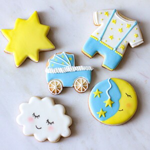 Puede incluir: Cinco galletas de az&uacute;car decoradas con temas de beb&eacute;. Una estrella amarilla, un mameluco de beb&eacute; azul con estrellas amarillas, un cochecito azul, una nube blanca con una cara dormida y una luna creciente amarilla con una cara dormida y tres estrellas amarillas.