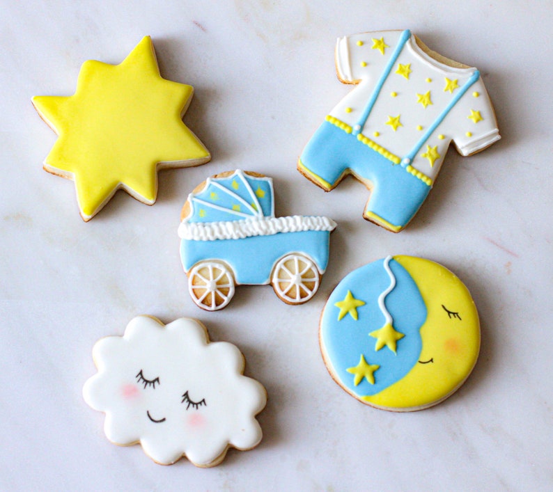 Puede incluir: Cinco galletas de az&uacute;car decoradas con temas de beb&eacute;. Una estrella amarilla, un mameluco de beb&eacute; azul con estrellas amarillas, un cochecito azul, una nube blanca con una cara dormida y una luna creciente amarilla con una cara dormida y tres estrellas amarillas.