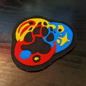 May include: A black fabric patch with a red, yellow, and blue paw print design. The paw print is surrounded by a red, yellow, and blue swirl.