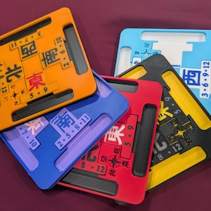 May include: A set of five square Mahjong wind indicators in orange, blue, red, yellow, and light blue. Each plastic tile features a central Chinese character for a cardinal direction, a compass rose icon, and numerical sequences printed in black or white.