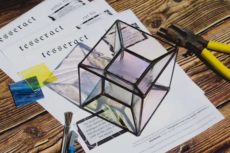 K&ouml;nnte beinhalten: Ein klarer Glas-Tesserakt, eine vierdimensionale geometrische Form, steht auf einer Holzoberfl&auml;che. Der Tesserakt besteht aus Buntglas und hat einen schwarzen Metallrahmen. In der N&auml;he befinden sich Werkzeuge und Buntglasst&uuml;cke, was auf ein Bastelprojekt hindeutet.