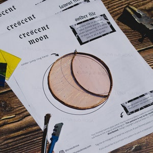 May include: A white paper template for a stained glass crescent moon project. The template includes instructions for soldering jump rings and a note about design credit. The image also shows a piece of stained glass, a pair of pliers, and a paintbrush.