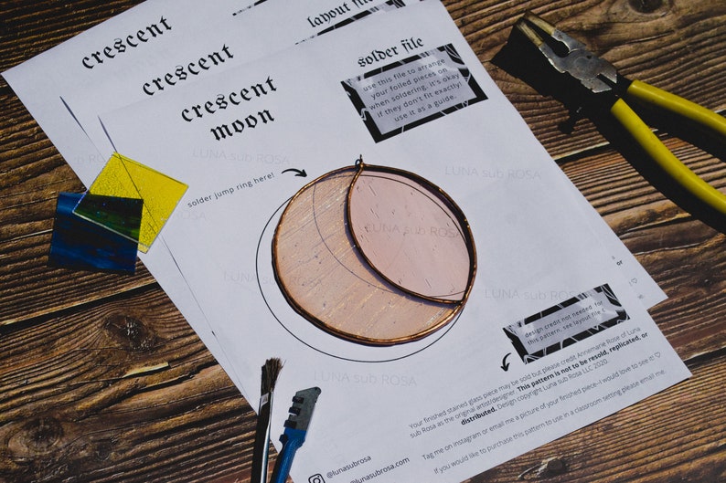 May include: A white paper template for a stained glass crescent moon project. The template includes instructions for soldering jump rings and a note about design credit. The image also shows a piece of stained glass, a pair of pliers, and a paintbrush.