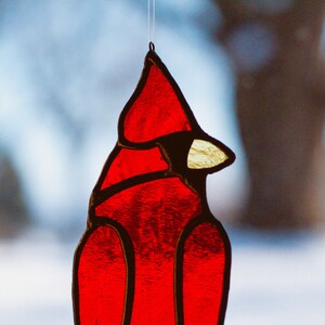 Op de afbeelding: Een rood glas-in-lood ornament van een kardinaal dat aan een raam hangt. De kardinaal is gedetailleerd met zwarte contouren en een gele snavel.